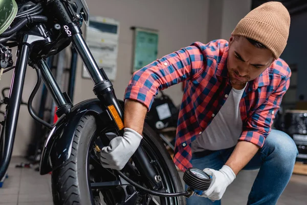 Mécanicien en chemise à carreaux et bonnet vérifier la pression d'air dans le pneu de la moto avec manomètre — Photo de stock