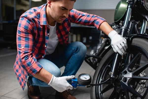 Young mechanic in plaid shirt measuring air pressure in tire of motorcycle with manometer — Stock Photo