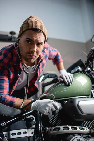 Mechanic in beanie and gloves looking at camera while examining engine of motorcycle with stethoscope — Stock Photo