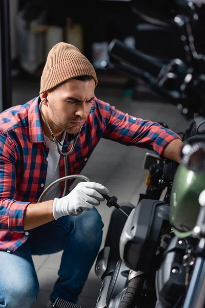 Mechanic in plaid shirt and gloves checking engine of motorcycle with stethoscope on blurred foreground — Stock Photo