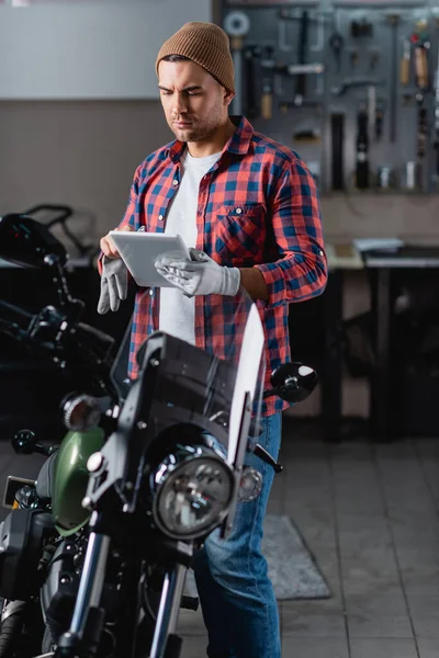 Молодой механик в шапочке и клетчатой рубашке с помощью цифрового планшета, стоя рядом с мотоциклом в мастерской — стоковое фото