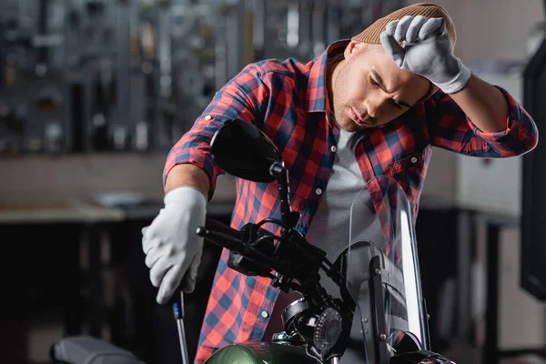 Tired mechanic holding screwdriver while examining motorcycle in workshop — Stock Photo