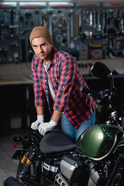 Jovem reparador em camisa xadrez e gorro olhando para a câmera enquanto estava perto de moto na oficina — Fotografia de Stock