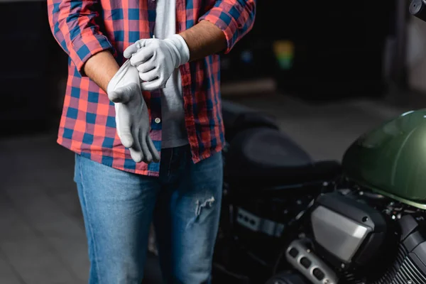 Vista recortada de mecánico en camisa a cuadros y jeans poniéndose guantes en taller - foto de stock