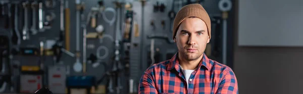 Young repairman in checkered shirt and beanie looking at camera in garage, banner — Stock Photo