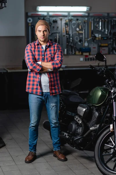 Visão de comprimento total do mecânico em camisa xadrez e jeans de pé com braços cruzados perto de motocicleta em oficina — Fotografia de Stock