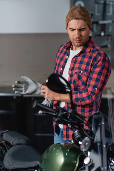 Joven mecánico en camisa a cuadros y gorro ponerse guantes de trabajo cerca de la motocicleta, borrosa primer plano - foto de stock