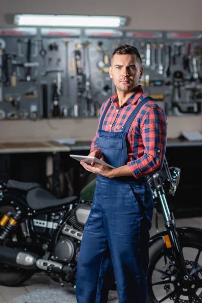 Joven técnico en overoles mirando a la cámara mientras está de pie cerca de la motocicleta con la tableta digital, fondo borroso - foto de stock