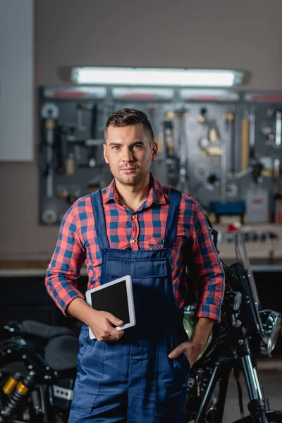 Mechanic in overalls with hand in pocket looking at camera near motorbike on blurred background — Stock Photo