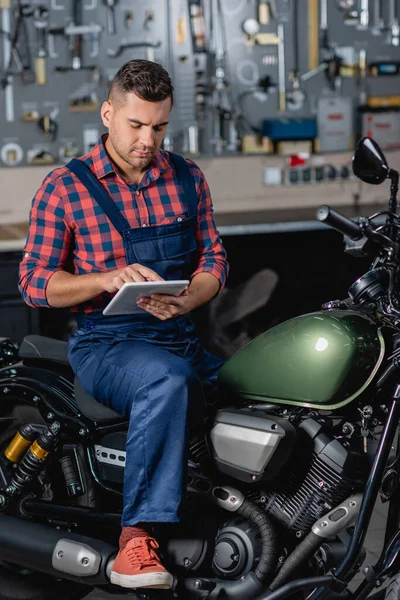 Meccanico in tuta da lavoro con tablet digitale seduto su moto in garage — Foto stock