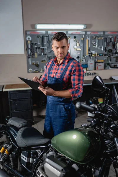 Técnico en overoles escribir en portapapeles cerca de la moto en el taller - foto de stock