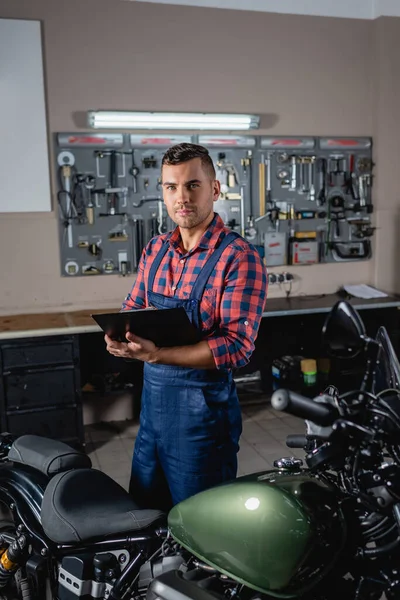 Joven técnico en overoles mirando a la cámara mientras está de pie con el portapapeles cerca de la motocicleta - foto de stock