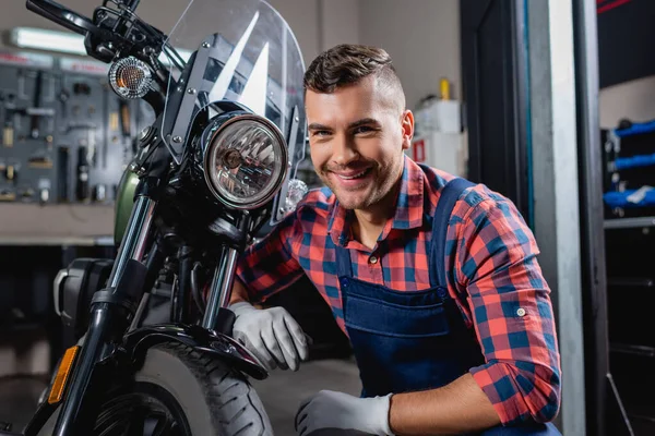 Joyeux mécanicien en salopette souriant à la caméra près de la moto en atelier — Photo de stock