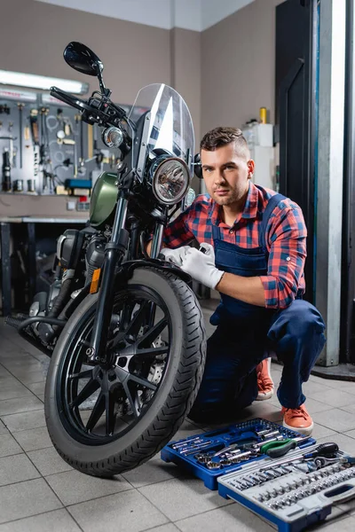 Joven técnico en overoles mirando la cámara cerca de la motocicleta y caja de herramientas en el taller - foto de stock