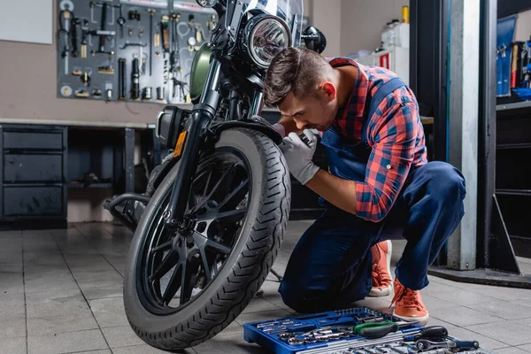 Joven técnico en overoles haciendo diagnósticos de motocicleta en garaje cerca de caja de herramientas - foto de stock