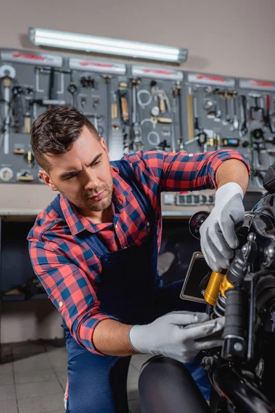 Jeune réparateur en chemise à carreaux et salopettes examen moto dans le garage — Photo de stock