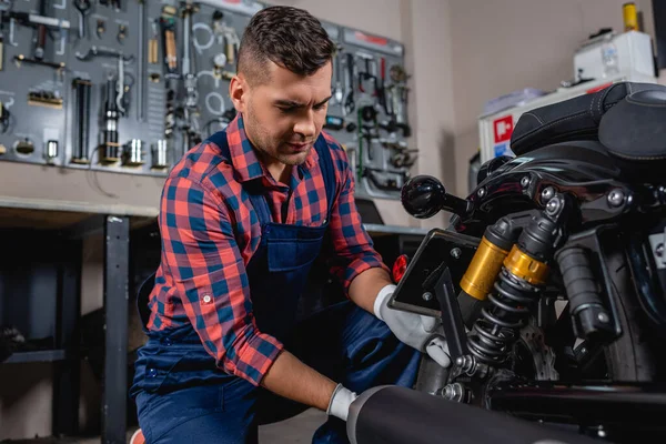 Jovem mecânico em macacão e camisa xadrez verificando amortecedor de motocicleta em oficina — Fotografia de Stock