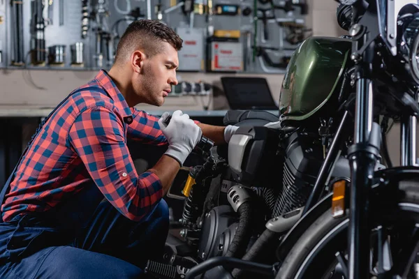 Mecánico en overoles usando linterna durante el diagnóstico de la motocicleta - foto de stock