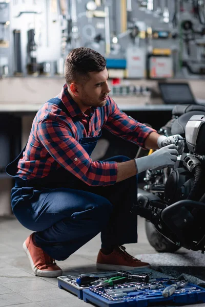 Joven mecánico en overoles haciendo diagnósticos de moto en taller cerca de caja de herramientas - foto de stock