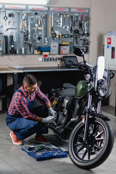 Mecánico en overoles haciendo diagnósticos de moto cerca de caja de herramientas en taller - foto de stock