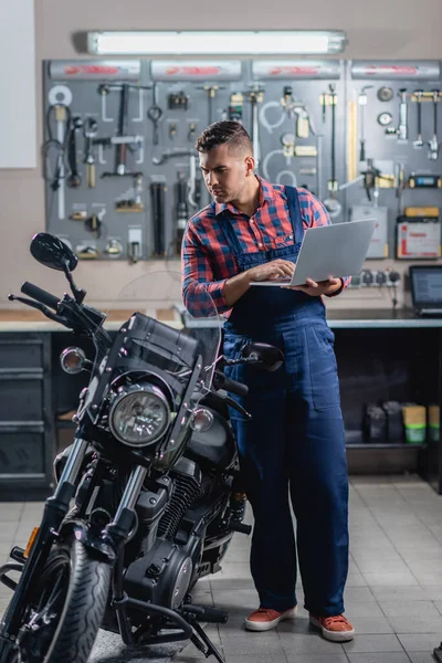 Vista integrale del meccanico in tuta da lavoro utilizzando laptop vicino alla moto in garage — Foto stock