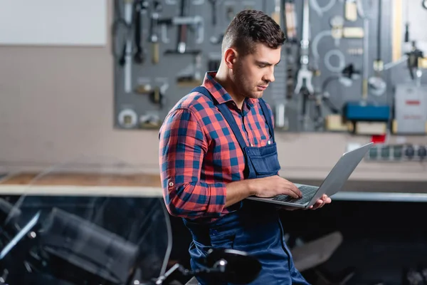 Young technician in overalls using laptop in workshop, blurred foreground — Stock Photo