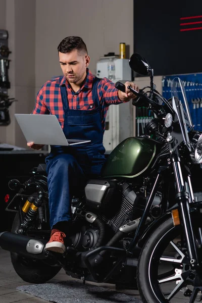 Jeune mécanicien en salopette à l'aide d'un ordinateur portable assis sur une moto en atelier — Photo de stock