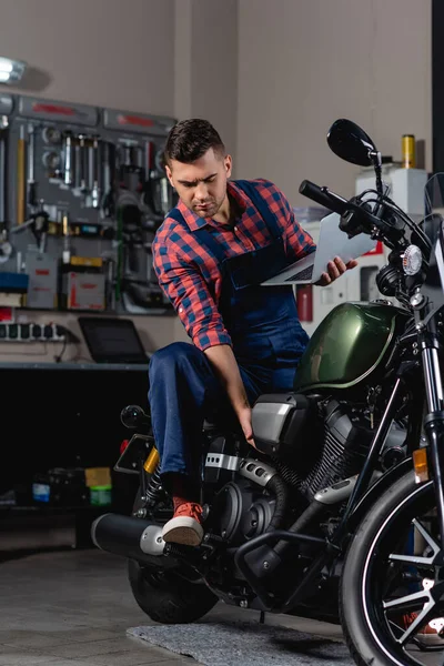 Reparador em macacão verificando motocicleta enquanto segurando laptop — Fotografia de Stock