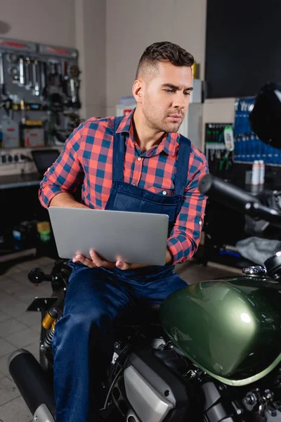 Young mechanic in overalls examining motorbike while holding laptop in workshop — Stock Photo