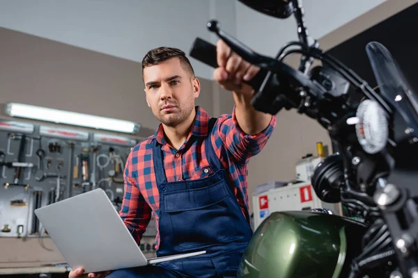 Junger Mechaniker mit Laptop blickt in Werkstatt auf Kamera, während er auf Motorrad sitzt, unscharfer Vordergrund — Stockfoto