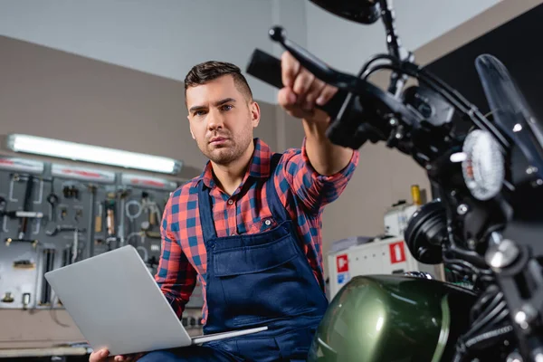 Giovane meccanico con computer portatile che guarda la macchina fotografica seduto sulla moto in officina, primo piano sfocato — Foto stock