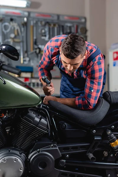 Jovem mecânico segurando chave de encaixe enquanto examina motocicleta na oficina — Fotografia de Stock