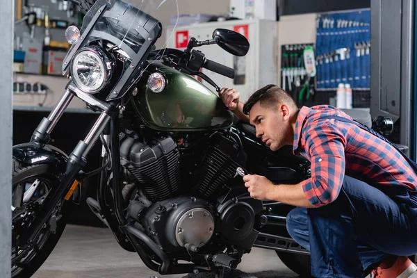 Jovem mecânico na camisa xadrez segurando chave de encaixe ao fazer diagnósticos de moto na oficina — Fotografia de Stock