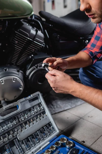 Vista recortada del reparador en guantes que sostienen la llave del zócalo cerca de la caja de herramientas y la motocicleta sobre fondo borroso - foto de stock