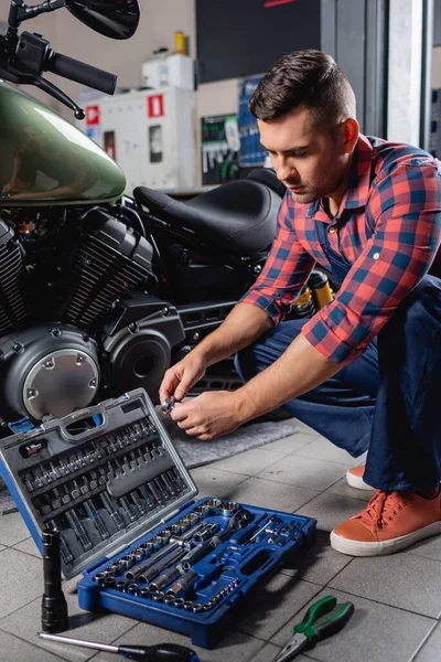 Joven técnico trabajando cerca de motocicleta y caja de herramientas en taller - foto de stock