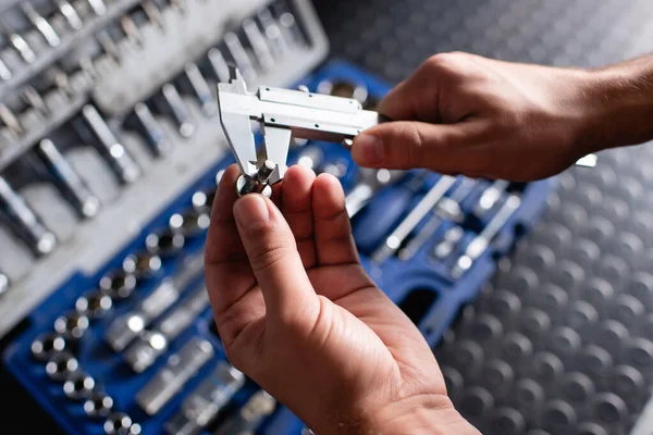 Cropped view of mechanic measuring size of srew with calipers near toolbox on blurred background — Stock Photo