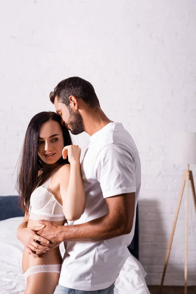 Улыбающийся молодой человек в футболке обнимающий женщину в нижнем белье в спальне — стоковое фото