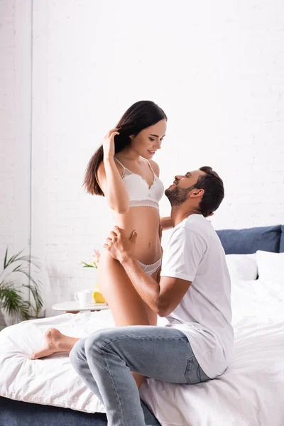 Smiling young man in t-shirt and jeans hugging and looking at seductive woman in underwear on bed — Stock Photo