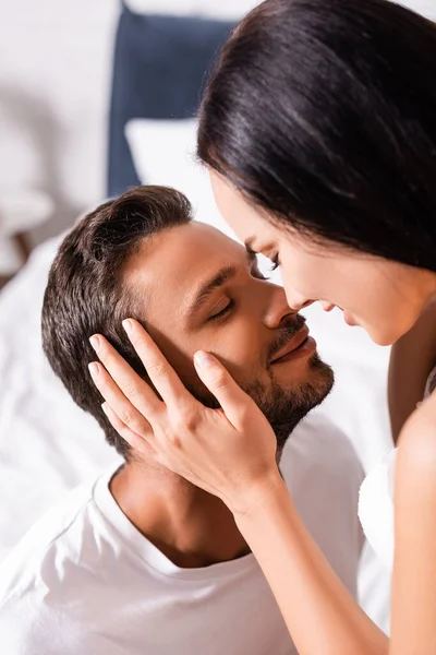 Happy brunette girlfriend holding hand on cheek of boyfriend with closed eyes on blurred background in bedroom — Stock Photo