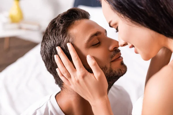 Sorridente ragazza bruna che tiene la mano sulla guancia del fidanzato con gli occhi chiusi su sfondo sfocato in camera da letto — Foto stock