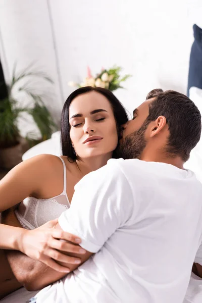 Young man hugging and kissing sensual brunette woman with closed eyes in bedroom on blurred background — Stock Photo