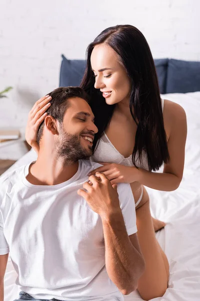 Feliz joven que se apoya en la mujer seductora en ropa interior de pie detrás de la cama - foto de stock