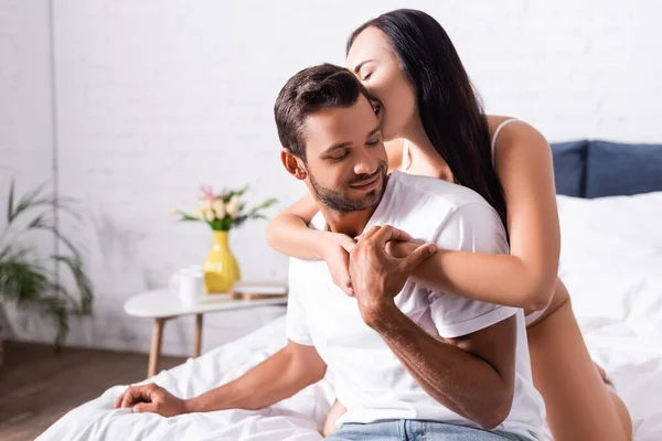 Seductora morena abrazando a un hombre sonriente con los ojos cerrados en el dormitorio - foto de stock