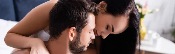 Спокуслива брюнетка жінка дивиться вниз, обіймаючи чоловіка на розмитому фоні в спальні, банер — стокове фото