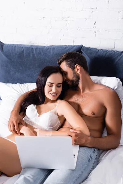Sonriente hombre sin camisa abrazando a la mujer en lencería en la cama cerca del ordenador portátil - foto de stock