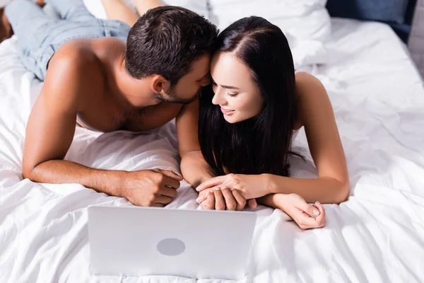 Sonriente sexy pareja cogida de la mano cerca de portátil en la cama sobre fondo borroso - foto de stock