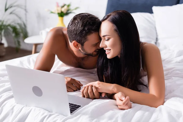 Glückliches Paar lacht, während es sich in der Nähe des Laptops auf verschwommenem Hintergrund im Schlafzimmer die Hände hält — Stockfoto