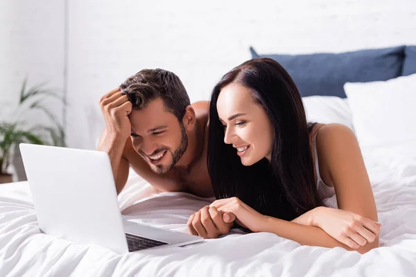 Feliz jovem casal rindo enquanto deitado na cama perto de laptop no fundo embaçado — Fotografia de Stock