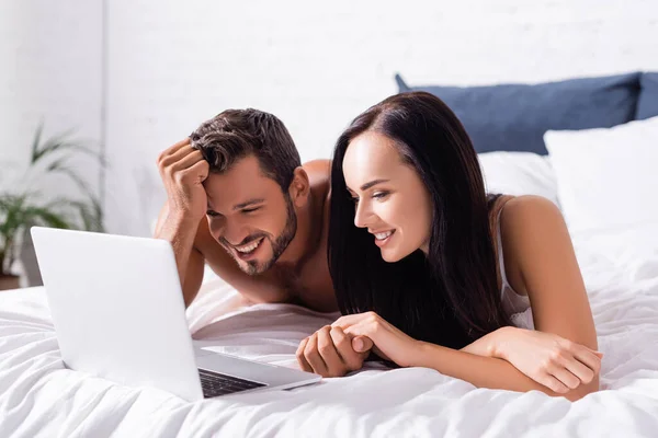 Glückliches junges Paar lacht, während es auf dem Bett in der Nähe von Laptop auf verschwommenem Hintergrund liegt — Stockfoto