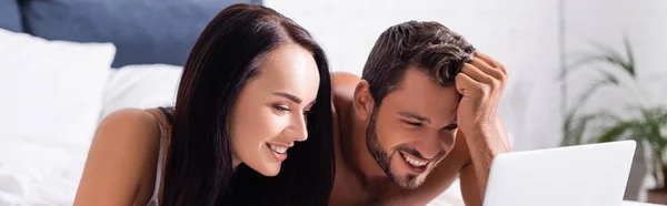 Feliz jovem casal rindo enquanto olha para laptop na cama no fundo borrado, banner — Fotografia de Stock